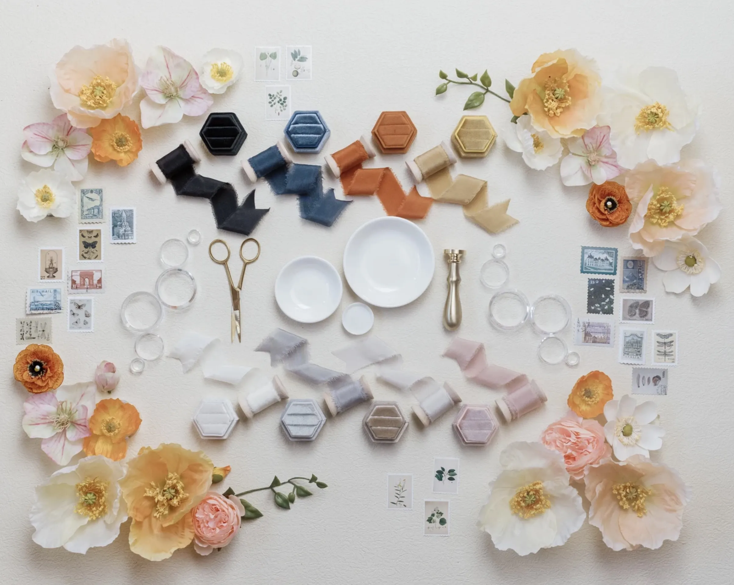 Overhead view of a wedding flat lay styling kit spread on a surface, with a velvet ring box, silk ribbon on a wooden spool, white styling dishes, acrylic blocks, vintage stamps and keys, gold scissors, and greenery arranged around an invitation suite